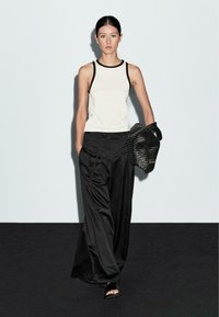 White tank top with black trim, paired with wide black trousers. Model holds a textured black woven bag; smooth flooring and light backdrop.