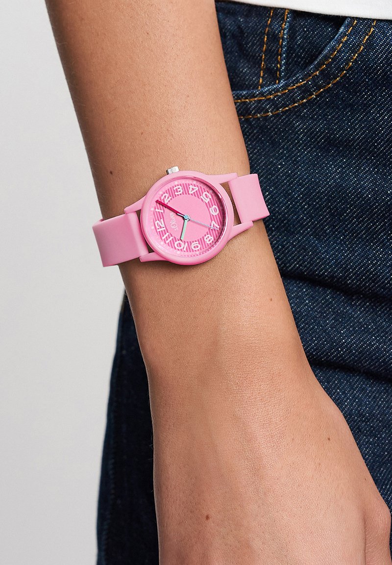 Poignet avec montre à bracelet en silicone rose affichant des chiffres blancs et un motif de cœur sur un cadran rose, portée avec un jean en denim bleu.