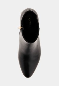 Stivale alla caviglia in pelle nera con punta affusolata e superficie liscia. Presenta un accento in oro con zip sul lato. All'interno è impresso "GUESS."