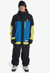 Skijack in zwart, blauw en neon-geel, met een ritssluiting op de borst, een capuchon en elastische manchetten, gecombineerd met losse zwarte broeken en handschoenen.
