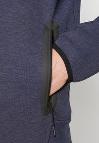 Marinblå sweatshirt med en dragkedjeficka på ärmen, med svart kant och en texturerad tyg. Hand placerad i fickan.