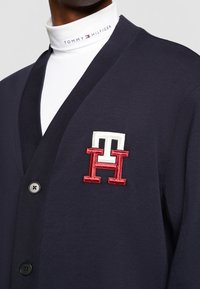 Cardigan blu navy con scollo a V, caratterizzato da un logo ricamato rosso e bianco sul petto e chiusura con bottoni. Realizzato in tessuto liscio.