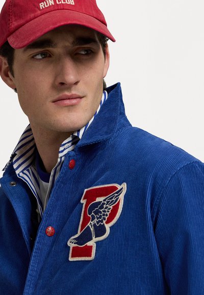 Blue corduroy jacket with a red and white embroidered "P" logo and wing detail; red snap buttons; layered with a striped shirt underneath.