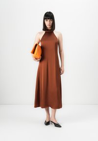 Kvinne med rett mørkt hår iført en ermeløs rustfarget kjole, holder en oransje clutch og har på seg svarte høyhælte sko, står mot en hvit bakgrunn.