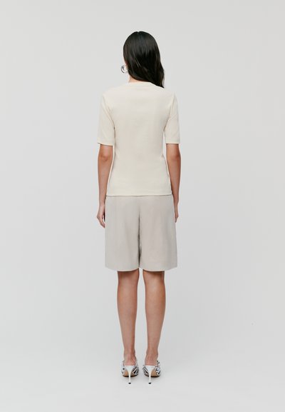 Krémový top s krátkym rukávom a priliehajúcim strihom, kombinovaný so svetlo šedými šortkami. Modelka má na sebe vzorované lodičky. Jednoduchý, minimalistický štýl.