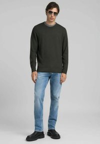 Olivengroene gebreide trui, blauwe spijkerbroek en zwarte stevige laarzen. Model draagt ook donkere zonnebril en staat voor een grijze achtergrond.