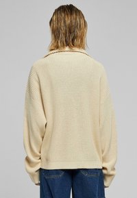 Béžový pletený svetr se širokým límcem, žebrovanou strukturou a spadlými rameny, kombinovaný s tmavými džínami. Pohled zezadu s jemným splýváním.