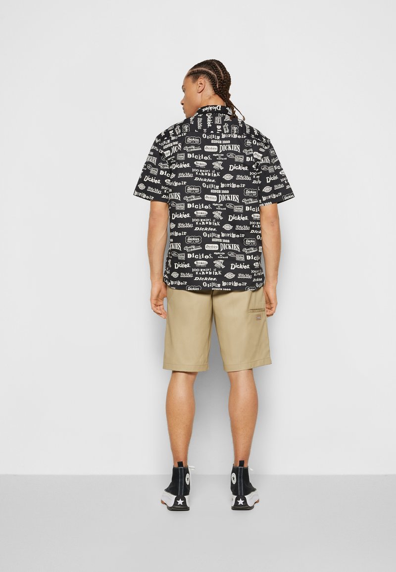 Black short-sleeve shirt with various logo prints, paired with tan shorts, and black high-top sneakers. Featuring a relaxed fit and visible stitching.