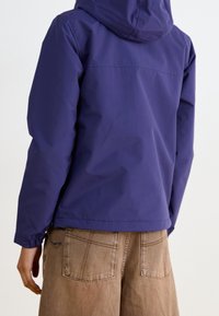 Persona vestida con una chaqueta con capucha púrpura y pantalones sueltos marrones, fotografiada de espaldas contra un fondo claro y liso.