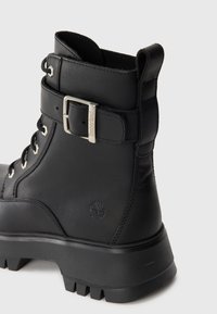 Čierne kožené poltopánky na šnurovanie s hrubou podrážkou, kotníkový remienok so striebornou sponou s vyrytým nápisom "Timberland" a embosovaným logom stromu na boku.