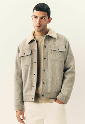 Gray wool jacket featuring a beige faux fur collar, two chest pockets, and snap buttons, worn over a beige shirt and light-colored trousers.