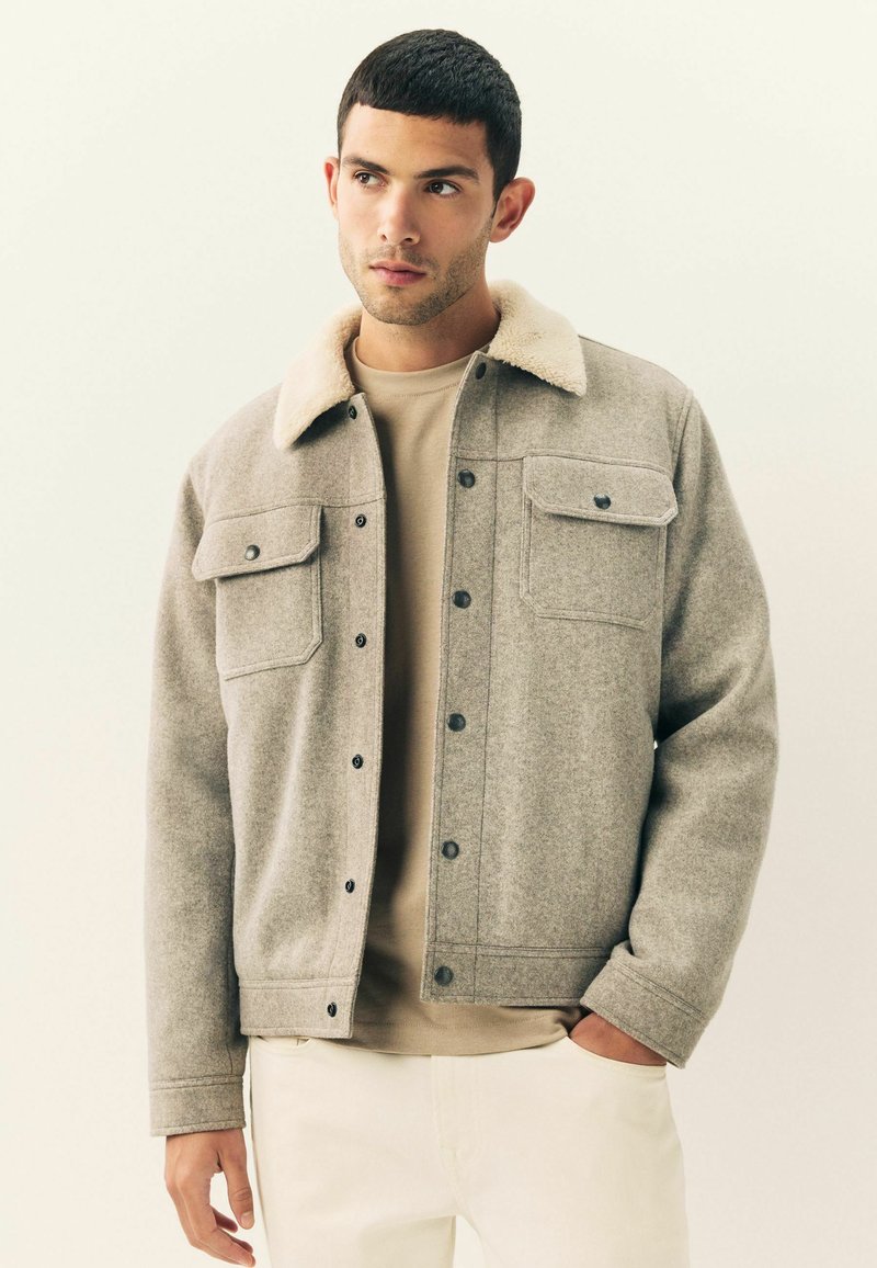 Grå uldjakke med en beige kunstpelskrave, to brystlommer og trykknapper, båret over en beige skjorte og lysfarvede bukser.