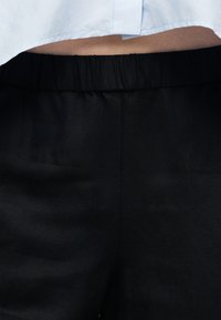 Pantalon noir à taille élastique porté avec un haut court blanc, laissant apparaître une petite partie du ventre.