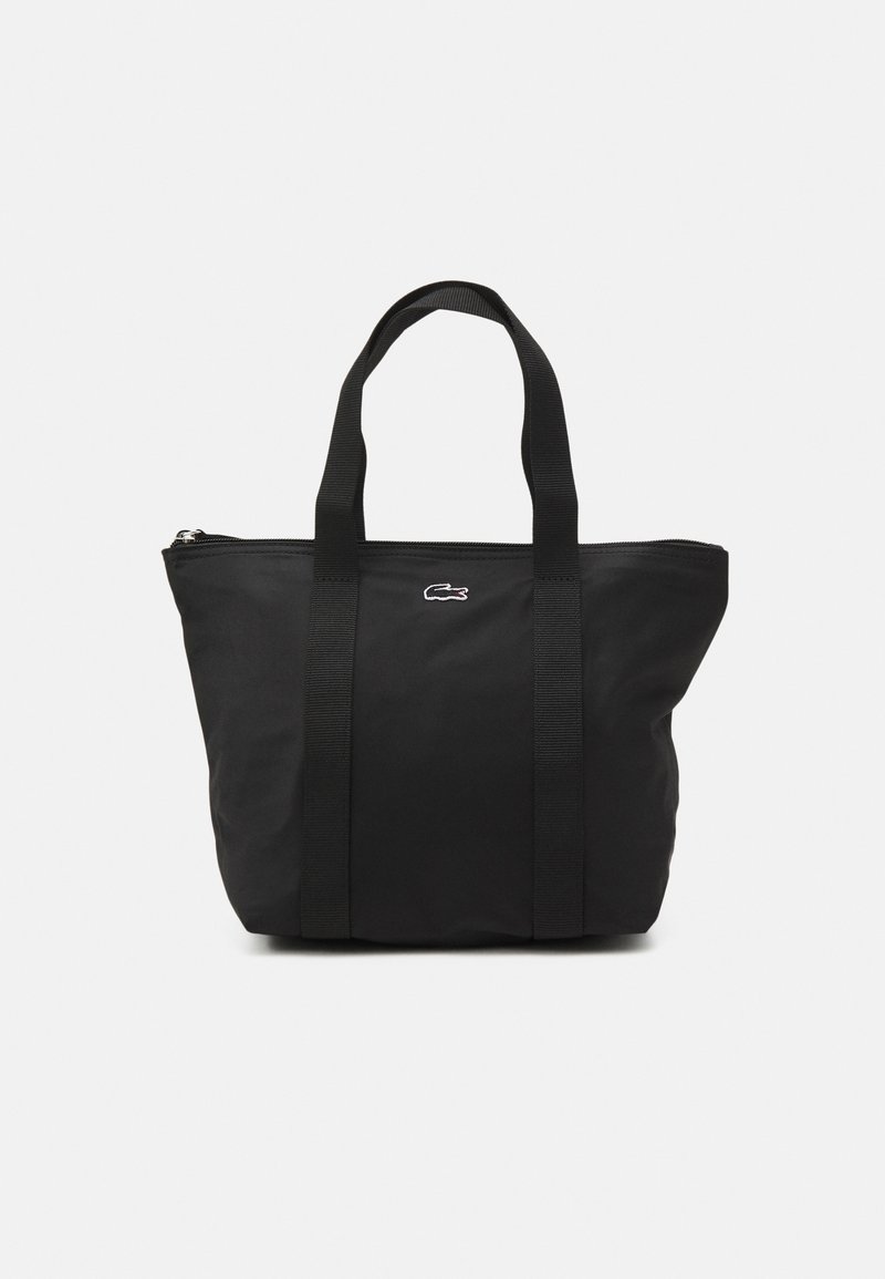 Sac cabas noir avec doubles poignées, fermeture éclair et petit logo crocodile blanc au centre avant sur fond gris clair.