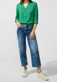 Grön knäppblus med v-ringning och uppvikta ärmar, ihopparad med ljusblå vida jeans och vita sneakers. Mjuk tyg och avslappnad passform.