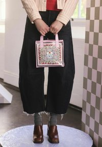 Modell hält eine rosa, dekorative Handtasche mit Blumen- und Spielkartenmotiven, trägt weite, dunkle Jeans und braune Plateauschuhe.