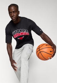 T-shirt nera a maniche corte con grafiche "CHICAGO" e logo del toro rosso, abbinata a pantaloni cargo grigi chiari. Modello che tiene in mano un pallone da basket arancione.