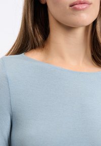 Gros plan du bas du visage d'une femme, de son cou et de son épaule, portant un haut bleu clair à encolure bateau et ayant de longs cheveux bruns.