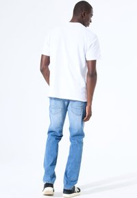 Hombre con camiseta blanca lisa, vaqueros de un azul claro y zapatillas negras, de pie de espaldas contra un fondo blanco.