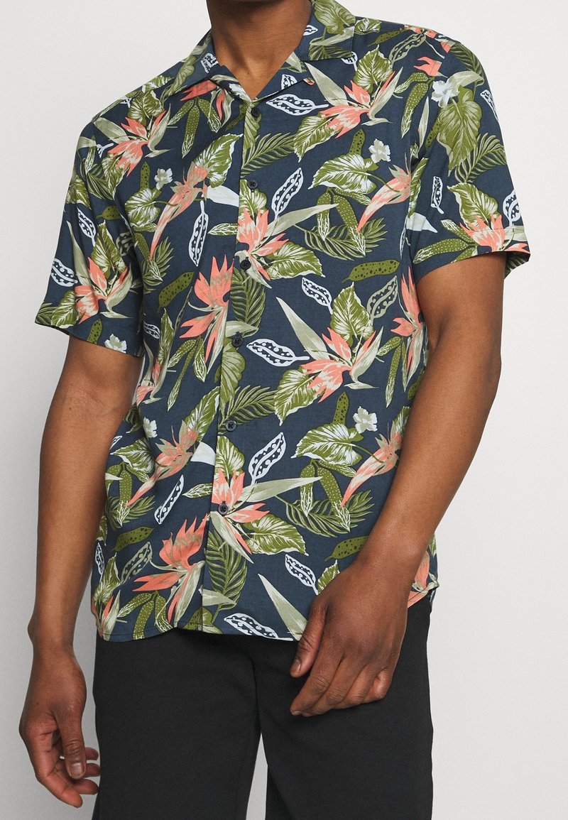 Chemise à manches courtes boutonnée avec fond bleu marine et motif de feuilles et fleurs tropicales vert, blanc et rose, portée avec un pantalon noir.