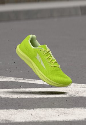 Zapato deportivo verde brillante con tela de malla y cordones, suspendido en el aire sobre una carretera de asfalto con marcas blancas, se pueden ver pequeños escombros.