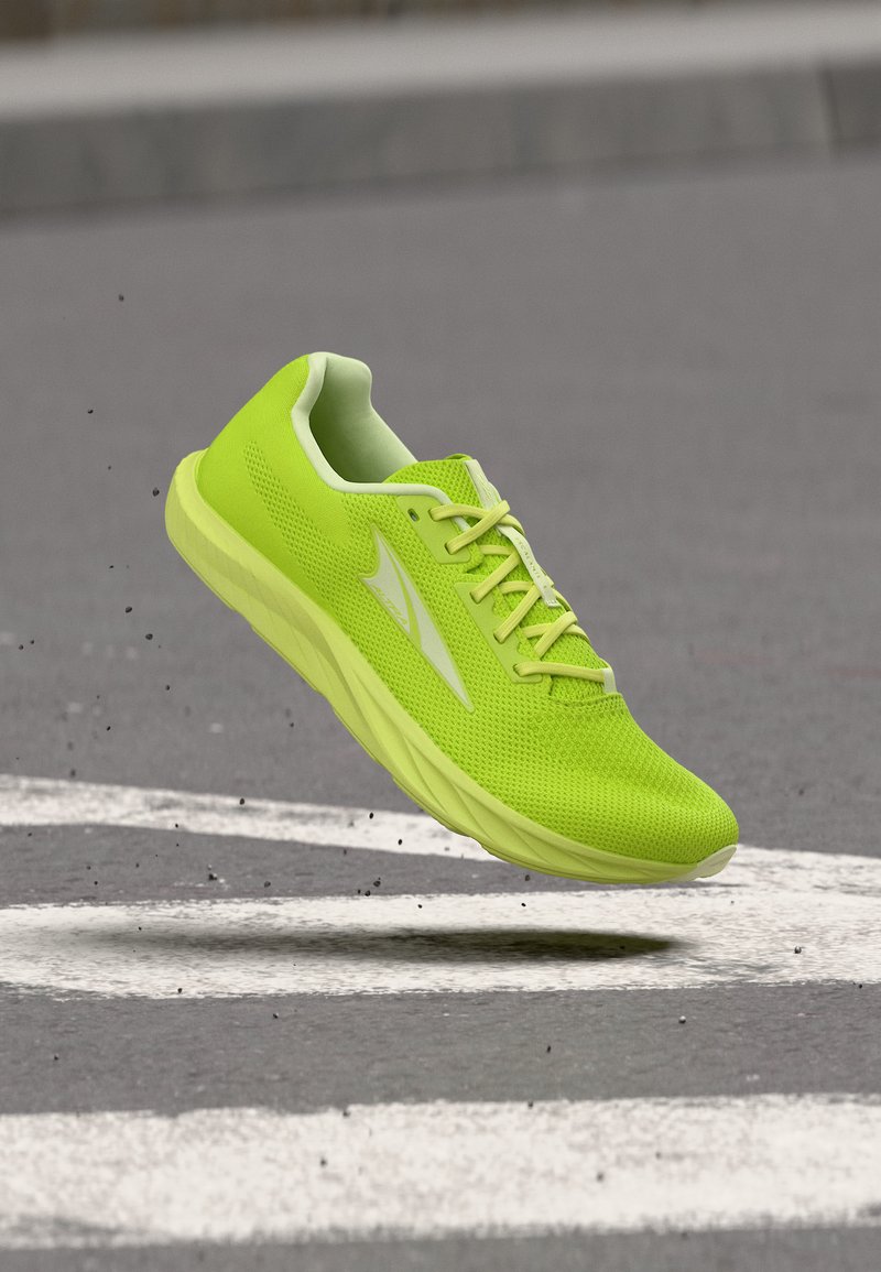 Jasně zelená sportovní obuv z síťoviny se šněrováním, ve vzduchu nad asfaltovou silnicí s bílými značkami, viditelné malé úlomky.