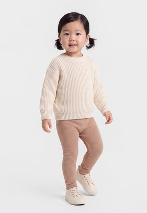 Lille barn med mørkt hår i fletninger iført en cremefarvet strikket sweater, lysebrune leggings og off-white sneakers står på en ensfarvet hvid baggrund.
