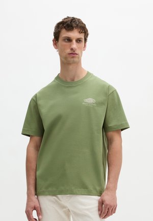 Młody mężczyzna z krótkimi kręconymi brązowymi włosami nosi zielony t-shirt z krótkim rękawem z małym logo na piersi oraz jasnobeżowe spodnie.