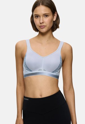 Jeune femme portant un soutien-gorge de sport gris clair avec des côtés en mesh à motifs et un legging noir taille haute sur un fond uni.