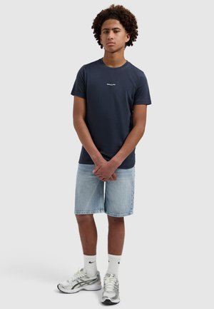 Jonge man met krullend haar, gekleed in een marineblauw "BALLIN" T-shirt, lichte denimshorts, witte Nike sokken en zilveren sportschoenen, staand met de handen gevouwen.