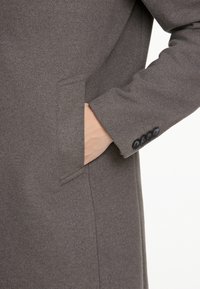 Cappotto grigio in lana con tasca laterale. La manica presenta un dettaglio con quattro bottoni al polso, conferendo un aspetto strutturato e sartoriale.