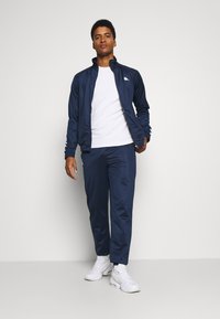 Férfi sportmelegítő sötét nápolyi színben, sima anyagból készült. A dzseki magas gallérral és fehér logóval rendelkezik; a nadrág rugalmas derékkal van ellátva.