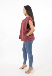 Burgunderroter Faux-Fur-Weste mit Seitentaschen, ärmellosem Design und lockerer Passform, kombiniert mit blauen Skinny-Jeans und klaren Absatzsandalen.