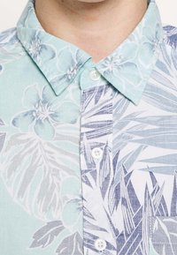 Koszula w jasnoniebieskie i białe kwiatowe wzory z kołnierzem typu spread, zapięciem na guziki i kontrastującymi wzorami liści i kwiatów. Miękka w dotyku.