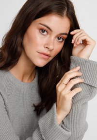 Un pull en tricot gris avec des poignets côtelés, porté par une personne affichant une fine bague en or à son doigt. Maquillage naturel et longs cheveux noirs visibles.