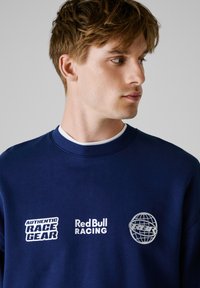 Sweatshirt bleu marine avec col rond côtelé, ornée de texte blanc "AUTHENTIC RACE GEAR" et "Red Bull RACING", ainsi que d'un logo globe argenté.