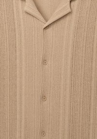 Beige gombos ing, függőleges csíkos és finom mintázatú texturált kötött anyagból. Klasszikus gallérral és öt gombbal rendelkezik.
