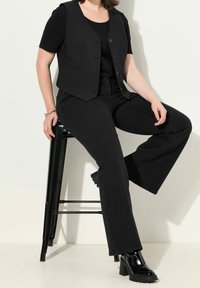 Schwarze Weste über einem schwarzen Oberteil, kombiniert mit ausgestellten schwarzen Jeans. Das Outfit umfasst glänzende schwarze Absatzstiefel und ein minimalistisches Design.