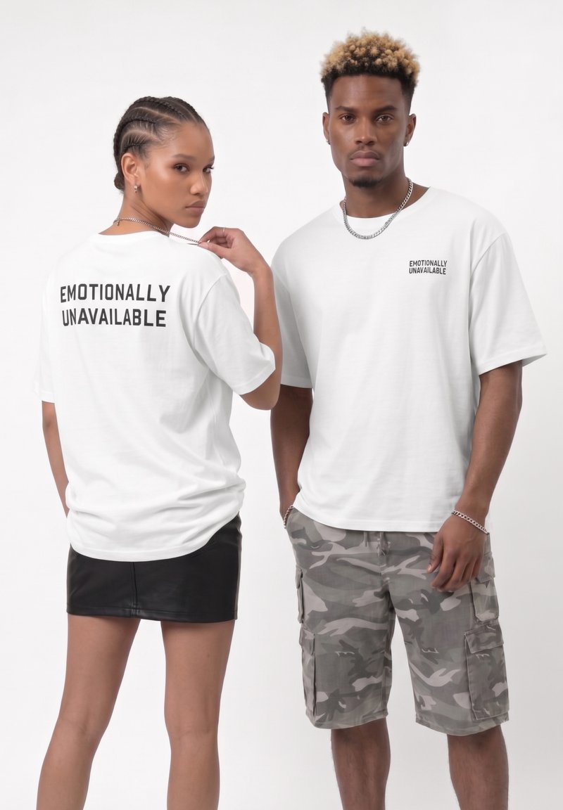 Δύο μοντέλα φορούν λευκά T-shirt με την επιγραφή «Emotionally Unavailable»· η γυναίκα έχει γυρισμένη την πλάτη, ο άντρας κοιτάει μπροστά, φοράει καμουφλάζ σορτς και μαύρη φούστα.