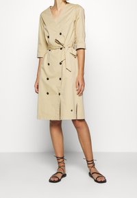 Sandelbeiges, knielanges Kleid mit Wickeldesign, V-Ausschnitt, dreiviertellangen Ärmeln, doppelter Knopfleiste, Taillengürtel und seitlichem Schlitz.