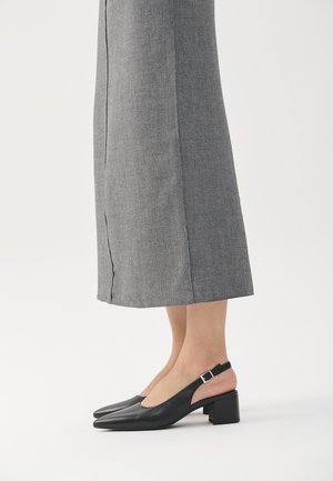 Beine in grauen, weit geschnittenen Hosen und schwarzen, ledernen Slingback-Schuhen mit niedrigem Blockabsatz und spitzen Zehen vor einem weißen Hintergrund.