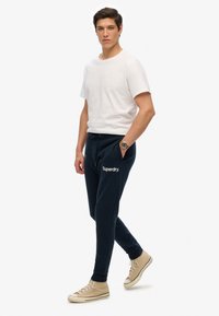 Ciemnoniebieskie spodnie dresowe z elastycznym pasem i sznurkiem, z białym logo "Superdry"; zestawione z prostą białą koszulką i beżowymi sneakersami.