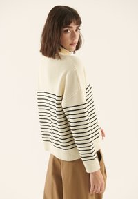 Adīts džemperis krēmkrāsā ar melnām horizontālām svītrām. Tam ir augsts apkaklis, nokarenas plecu līnijas un ribotu apmali piedurknēm un apakšmalai. Mīksta tekstūra.