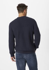 Marineblauer Strickpullover mit strukturierten Mustern im oberen Bereich und gerippten Bündchen. Kombiniert mit hellblauen Jeans.