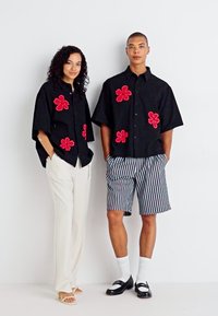 Sorte kortærmede skjorter med rød blomsterbroderi, parret med hvide bukser og stribede shorts. Accessories inkluderer sandaler og loafers.