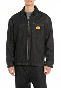 Zwarte denim zip-up jack met een puntkraag, een borstzak met een oranje logopatch en drukknopen op de manchet.