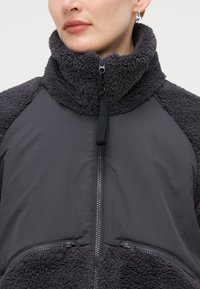 Zip-up jakna z debelim, puhast ovratnikom in rokavi v temno sivi barvi, z gladkim, lahkim telesom iz tkanine in z zadrgo zaprtimi žepi.