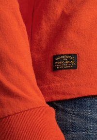 Maglione di cotone arancione con polsini a costine e un'etichetta nera con testo oro, parzialmente visibile su uno sfondo in denim blu.