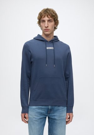Junger Mann mit blauem Calvin Klein Hoodie und hellblauen Jeans, stehend vor einem schlichten weißen Hintergrund.