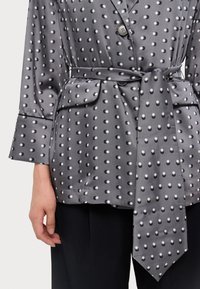 Bluza s točkicama od sive tkanine, s velikim bijelim točkicama. Ima ovratnik, kopčanje na dugmad i remen za vezanje u struku.